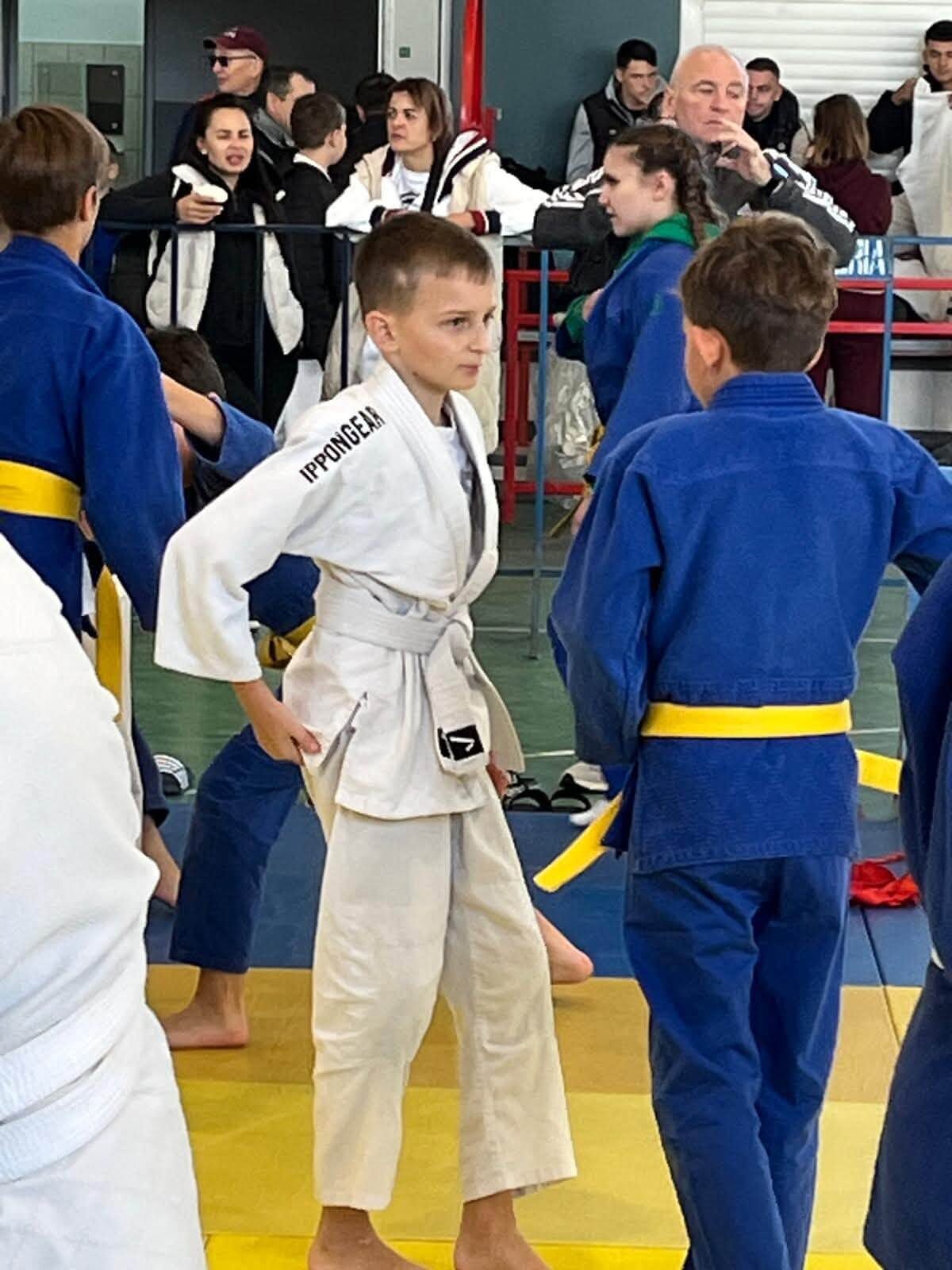 Tineri sportivi în kimono alb și albastru pregătindu-se lângă saltea, cu public în spate
